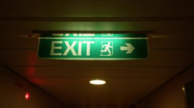 Işıklandırılmış yeşil çıkış işareti loş bir koridorda parlıyor. Binanın içindeki güvenlik yönünü ve acil durum yolunu gösteriyor.