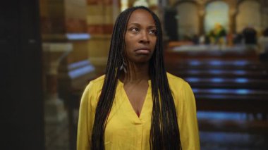 Arka planda sıra sıra sıralanmış, loş ışıklı bir kilisede sarı gömlekli bir kadın..