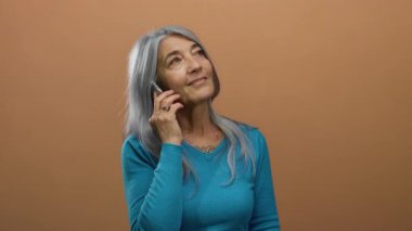 Gri saçlı yaşlı kadın kahverengi arka planda akıllı telefondan konuşuyor, yalnızken neşe ve iletişimi ifade ediyor..