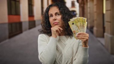 Bir şehir caddesinde Filipinli pezosu tutan kadın, finansal kararları derinden düşünüyor, şehir çevresi ile çevrili, orta yaşlarda merak ve düşünce ifade ediyor.