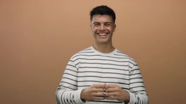 Genç adam gülümserken elleri kahverengi arka planda, çizgili bir gömlek giymiş, basit bir portrede mutluluğu ve sıcaklığı taşıyan bir kalp şekli yaratıyor..