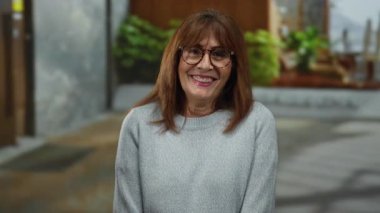 Kıdemli İspanyol kadın neşeyle gülümsüyor güneşli bir şehir caddesinde çevresi yeşillik ve modern binalarla çevrili şehir yaşam tarzını ve canlılığını yansıtan