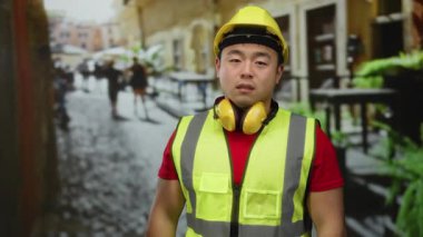 Şehir merkezinde arka planda bulanık duran, uykulu ve yorgun görünen, güvenlik yeleği ve sert şapka giyen bir adam çevredeki şehir hayatını gözler önüne seriyor..