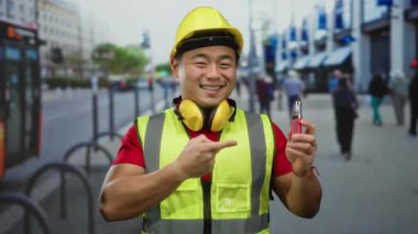 Çelik yelek giyen ve kasketli genç adam şehir arka planında bulanık şehir geçmişi olan ve yürüyen insanları hedef alan bir kalem taşıyor..