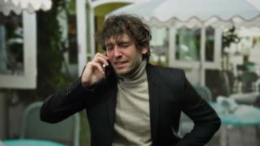 Dışarıda telefonla konuşan yakışıklı bir adam şehir terasında halka açık bir kafe ya da restorana benzeyen bir ceket giymekten rahatsız görünüyor..