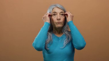 Gri saçlı, mavi kazaklı yaşlı kadın kahverengi arka planda izole edilmiş ellerini kaldırarak sürpriz yapıyor..