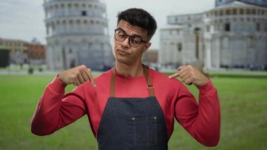 Genç adam gözlüklü ve önlüklü, açık hava sidikli sembolik kulesinin önünde jest yapıyor..
