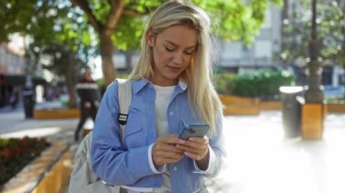 Şehir manzarası, ağaçlar ve doğa ile çevrili güneşli şehir parkında akıllı telefon kullanan bir kadın teknoloji ve sükunetin bir karışımını oluşturuyor..