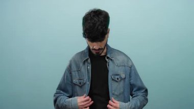Genç adam kot ceketini mavi arka plana göre ayarlıyor, şık ve düşünceli görünüyor, modern moda ve kendine güveni bir nebze de olsa gösteriyor..