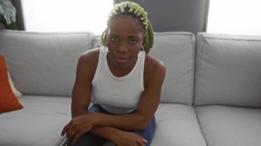 Örgülü kadın, şık oturma odasındaki koltuğa oturmuş kafaya dokunuyor. Parlak yastıklar içerideki modern yaşam tarzını gösteriyor..