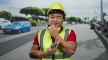 Emniyet yeleği ve miğfer giyen bir adam şehrin sokaklarında elinde nakit tutarak düşünceli duruyor, finans ve inşaat odaklı kentsel yaşam tarzı zorluklarını yansıtıyor..