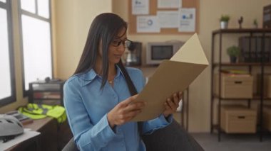 Mavi gömlekli genç bir kadın modern bir ofiste bir klasörü inceliyor, odak ve konsantrasyonunu, organize dekorasyon ve profesyonel kıyafetlerle kapalı bir ortamda gösteriyor..