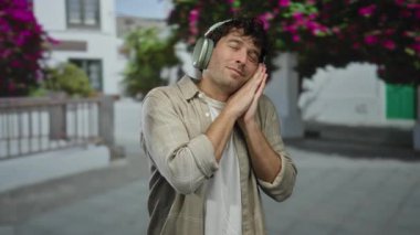 Kulaklık takan İspanyol bir adam uyuma hareketi yapıyor, çiçeklerle dolu bir sokakta duruyor, sakinliğini ve müziğin tadını çıkarıyor..