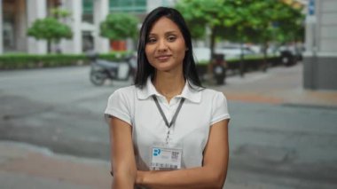 Arka planda binalar ve ağaçlarla dolu bir sokakta güvenle duran genç bir kadın, beyaz bir gömlek ve isim kartı takıyor, profesyonel bir bağlam olduğunu gösteriyor.