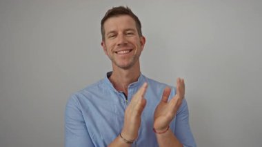 Mavi gömlekli bir adam beyaz bir arkaplanda gülümsüyor ve alkışlıyor, pozitifliğini ve neşesini izole ve sıradan bir ortamda ifade ediyor, modern moda anlayışını sergiliyor..