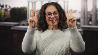 Kıvırcık saçlı, gülümseyen ve el hareketi yapan bir kadın şehir merkezinde neşeli bir atmosfer sergileyen binaların bulanık arka planına sahip..