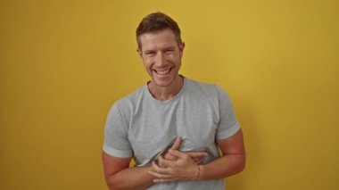 Genç bir adam, parlak sarı bir arka plana karşı güvenle gülümsüyor, günlük gri bir gömlek giyiyor, neşeli ve yakışıklı bir tavır sergiliyor..