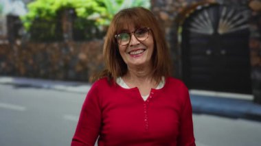 Kırmızı kazaklı son sınıf öğrencisi bir kadın, gülümsüyor ve şehir dışında bir sokakta başparmağıyla selam veriyor. Şehir manzarasında sıcaklık ve dostluk yayıyor..