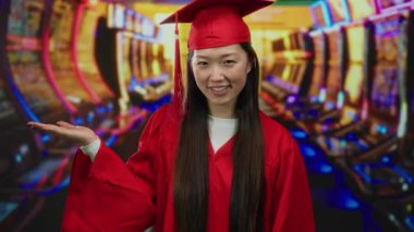 Kumarhanede mezuniyet elbisesi giymiş arka planda kumar makineleri olan bir kadın, gülümseyerek başarısını kutluyor, heyecan verici bir iç mekanda neşe ve başarı sergiliyor..