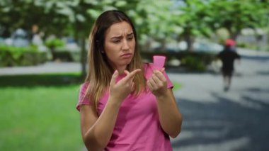 Parkta elinde regl fincanı tutan bir kadın, pembe tişörtlü, düşünceli bir yüz ifadesi olan, yeşil bulanık arka planda duran, çevre dostu yaşam tarzı tercihlerini vurgulayan..