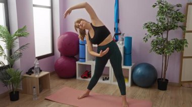 Spor salonunda yoga minderinde egzersiz yapan bir kadın canlı fitness ortamında esneklik gösteriyor. Buna pilates topları ve canlı atmosferi geliştiren saksı bitkileri de dahil..