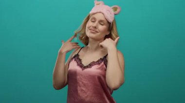 Pembe gecelikli ve uyku maskeli kadın canlı mavi bir arka plana karşı güvenle poz veriyor..