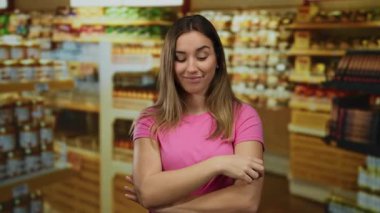 Genç, sarışın bir kadın, süpermarketin koridorunda kendine güvenen pembe bir gömlek giyiyor. Etrafı market eşyalarıyla çevrili.