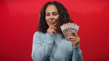 Meksika pesolarını elinde tutan kadın canlı kırmızı bir arkaplan karşısında gülümsüyor, zenginliği ve finansal temayı sembolize ediyor.