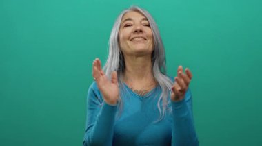 Gri saçlı yaşlı bir kadın yeşil arka plan duvarına karşı alkışlıyor, sıcacık mavi elbisesiyle gülümsüyor, neşe ve takdiri gösteriyor..