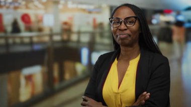 Alışveriş merkezinde gülümseyen bir kadın arka planda bulanık bir şekilde mağazanın ışıklarını ve canlı renklerini gösteriyor, kendine güven ve tarz yayıyor ama profesyonel kıyafetiyle.