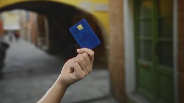 Elinde mavi bir kredi kartı olan beyaz bir adamın elinde yeşil kapılı ve arka planda kemerli bir caddede, şehir içi mali faaliyetlerini aktarıyor..