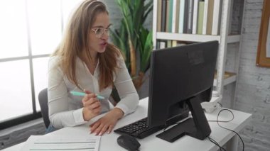 Ofiste çalışan bir kadın elinde kalemle bilgisayar başında kitap rafları ve arka planda görünür bitkiler olan modern bir ortamda kalem tutuyor.