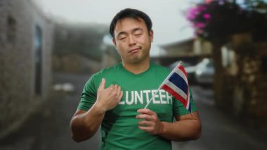 Sokaklarda Tayland bayrağı taşıyan genç adam gönüllü tişört giyiyor. Şehir ortamında kamu hizmeti öneriyor.