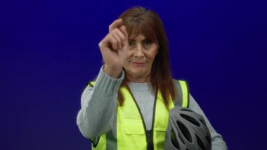 Güvenlik yeleği giyen, mavi duvara karşı kaskı tutan, güven ve profesyonellik taşıyan, ileri doğru gülümseyen bir kadın.