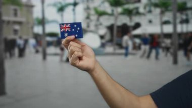 Açık şehir sokak ortamında nakışlı bir Avustralya bayrağı taşıyan bir adam arka planda insanların olduğu canlı bir şehir manzarası sergiliyor..