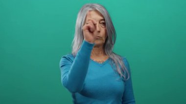 Gri saçlı yaşlı kadın, canlı yeşil bir arka plana karşı küçümseyici bir jest yapıyor ve güçlü bir kınama mesajı gönderiyor..