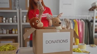 Yardım odasında içi doldurulmuş hayvanlarla bağış organize eden, gönüllü tişörtü giyen, telefonla konuşan ev ortamında giysi ve malzemelerle çevrili bir kadın..