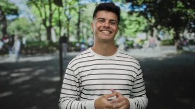 Çizgili kazaklı genç adam güneşli bir parkta arka planda bulanık insanlar ve ağaçlarla iyi bir jest yapıyor..