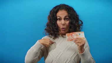 Orta yaşlı bir kadın, mavi bir arka plan duvarına karşı Filipinli bir peso banknotu tutarak, duygu ve merak göstererek kendisini işaret ediyor..