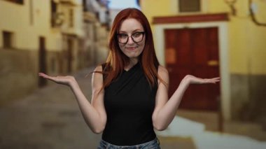 Kızıl saçlı ve gözlüklü bir kadın şehir caddesinde duruyor. Bulanık binalar, siyah bir bluz ve kot pantolon giyiyor. İki eliyle sorgulama jesti yapıyor..