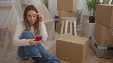 Akıllı telefonuyla yerde oturan bir kadın yeni evdeki kutuları taşıyarak genç ve rahat bir atmosferi canlandırıyor..