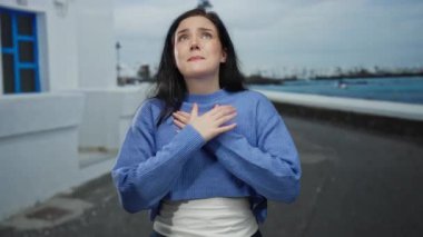 Sahilde duygularını ifade eden bir kadın mavi kazak giyiyor, yoğun duygular yakalıyor, kumral saçlar, deniz kenarı kentlerinde kendini kucaklıyor, sakin ama duygusal bir sahne.