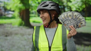 Başlık ve güvenlik yeleği giyen yaşlı kadın Amerikan dolarlarını parkın dışında tutuyor, mutluluk ve başarı yayıyor..