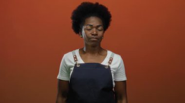 Önlük giyen kadın sakin bir şekilde kırmızı arka plana karşı duruyor gözleri kapalı ve sakin bir ifadeyle, canlı arkaplana karşı basit bir moda sergiliyor..