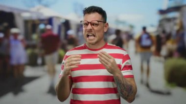 Çizgili tişörtlü ve dövmeli bir adam kalabalık bir şehir sokağında açık mavi gökyüzü altında hareketli hareketler yapıyor..