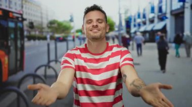 İşlek bir şehir sokağında dövmeleri ve çizgili gömleği olan genç bir adam arka planda insanlar ve araçlarla açık şehir ortamının tadını çıkarıyor..