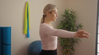 Spor salonunda egzersiz yapan, sarı saçlı, pembe tişörtlü, yoga minderleri, egzersiz topları ve kapalı alandaki bitkilerle dolu egzersiz rutinine odaklanan bir kadın..