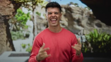 Şehir caddesinde kırmızı bir kazakla, arka planda ağaçlarla neşeli jestler yapan genç adam güneşli bir günde enerjik bir şekilde gülümsüyor..