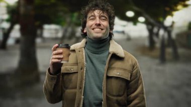Şehrin dışında elinde kahve tutan ve neşeli bir ifadeyle kameraya bakan genç adam, arka planda bulanık yeşil ağaçlar olan şehir sokaklarında ceket giyiyor..