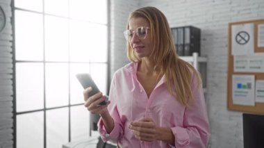 Pembe tişörtlü gülümseyen kadın telefonda konuşuyor. Geniş pencereli ve düzenli çalışma alanı olan parlak ofis ortamında profesyonel ve dostane bir atmosferi aktarıyor..